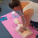 Learner performing hands‑only CPR compressions on a training manikin on a pink exercise mat.