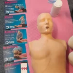 CPR training manikin placed beside step‑by‑step first aid instruction cards on a mat.
