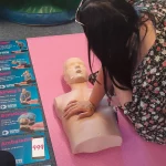 A trainee practicing life-saving techniques next to a series of instructional posters detailing the DRABC (Danger, Response, Airway, Breathing, Circulation) steps.