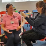 Two people in a first aid class demonstrating how to apply a bandage to an arm while seated at a table in a classroom setting.