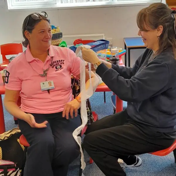 Two people in a first aid class demonstrating how to apply a bandage to an arm while seated at a table in a classroom setting.