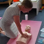 A woman kneeling on a mat practicing the correct hand placement and body posture for adult chest compressions on a manikin.