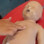 Close-up of the two-finger chest compression technique being practiced on an infant CPR training manikin.