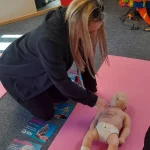 Person practising infant CPR compressions on a baby training manikin during a first aid session.